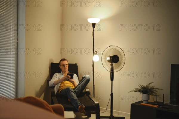 A man sits comfortably in a chair and reading a story on e-book while relaxing in a cozy living room. Soft lighting from a floor lamp and a fan creates a calm atmosphere during the evening