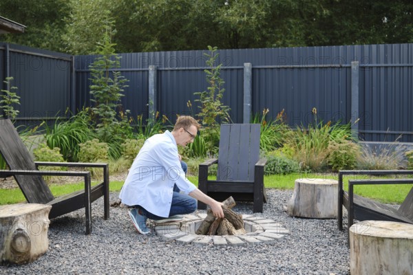A middle-aged man dressed in a light blue shirt arranges firewood at a circular stone fire pit surrounded by wooden seating