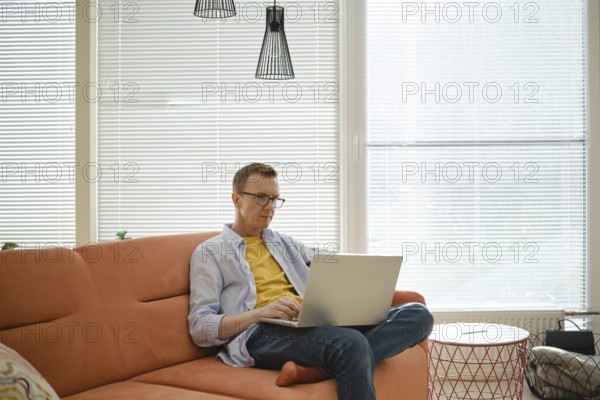A man is seated on an orange couch, focused on his work on a laptop. Sunlight streams through the window, illuminating modern decor in a cozy living space that emphasizes a relaxed atmosphere