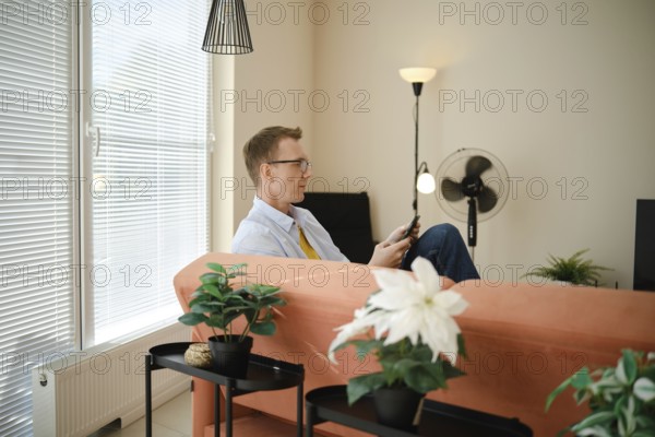 A man sits comfortably on a couch in a modern living room with large windows and natural light. He is holding a smartphone and surfing the web during peaceful afternoon
