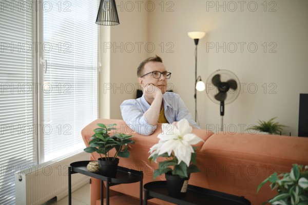 A man sits thoughtfully on a couch in a cozy living room, enjoying a moment of reflection. Bright natural light streams through large windows, illuminating indoor plants and stylish decor