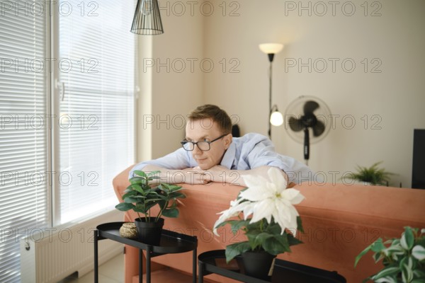 A middle-aged man leans on a colourful couch, gazing thoughtfully at potted indoor plants nearby. Sunlight pours through large windows, creating a serene atmosphere in the stylish living space