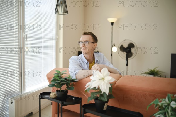 A middle-aged man sits comfortably on a stylish couch, looking contemplatively out of large windows. Sunlight streams into a contemporary living room adorned with potted plants and a ceiling fan