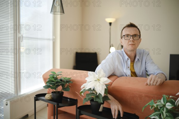 A middle-aged man sits on an orange couch in a contemporary living room. He appears thoughtful as he leans against the couch, surrounded by stylish potted plants and a warm ambiance