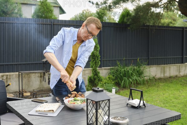 A man sprinkles fresh vegetables with salt in the backyard. Smoke from a barbecue can be seen in the background