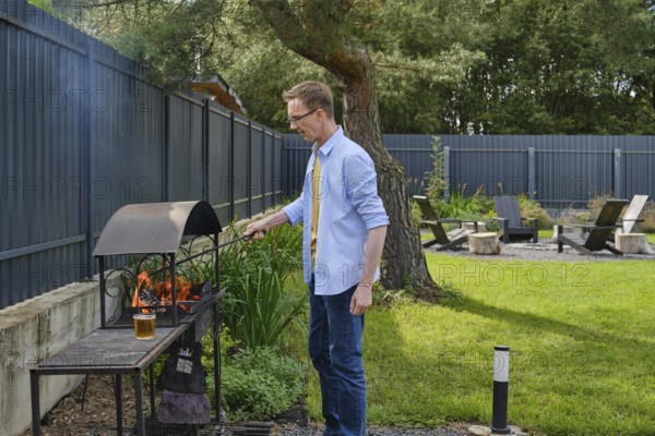 A man stands beside a barbecue grill, actively tending to the flames in a vibrant backyard. The scene shows lush greenery, outdoor seating, and a relaxed atmosphere under sunny skies