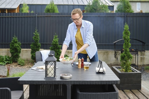 A man is arranging a fresh salad on a table in an inviting garden area. The sun is shining brightly, illuminating the surrounding greenery and modern outdoor furniture