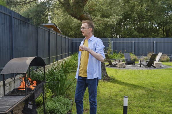 A man stands next to a barbecue grill, casually holding a drink while readying food. The scene is set in a lush garden with green grass, wooden seating, and a blue sky in the background
