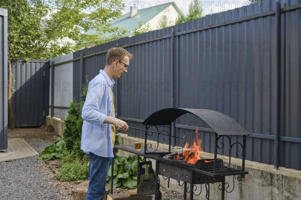 A man stands by a grill in a backyard, tending to the flames. The surrounding area features greenery and a fence, creating a cozy outdoor cooking environment
