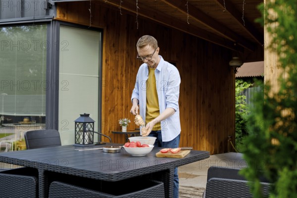A man is joyfully preparing a meal outdoors at a wooden kitchen area. He strands meat on a skewer enjoying the warm afternoon sunlight and inviting environment