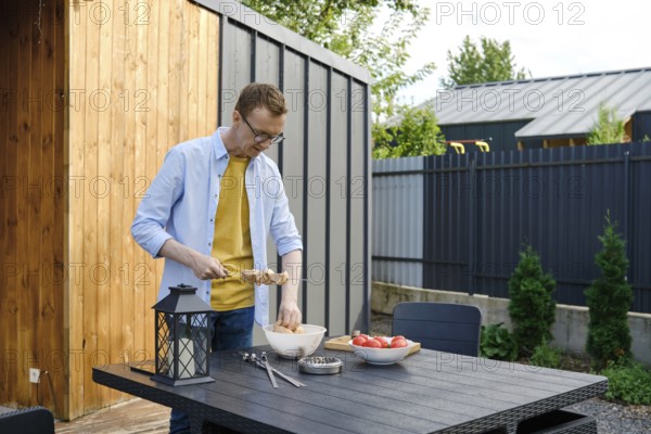A man wearing a yellow shirt and denim jacket is preparing meat skewers at a wooden table in a backyard. Fresh vegetables are on the table, and greenery surrounds the area during a sunny day