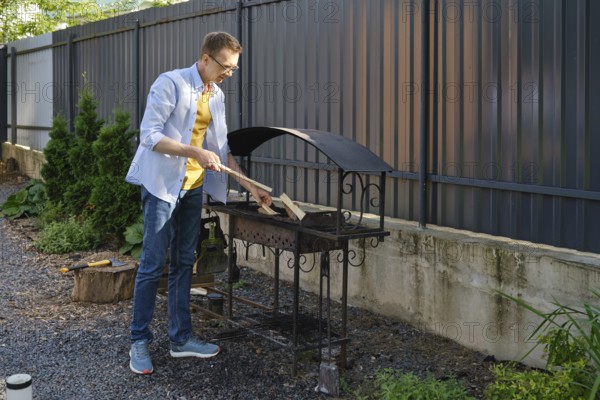 A middle-aged a man puts firewood in the grill in a backyard. He is going to cook meat outdoors on a sunny day