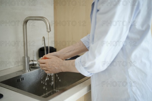 Hands gently scrub a fresh tomato under running water at a kitchen sink, emphasizing cleanliness during meal prep. The setting is a contemporary kitchen with simple, elegant design