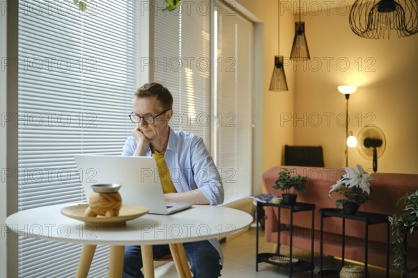 A man sits at a round table and watching sports on laptop, in a bright and airy living room. Sunlight filters through window blinds, illuminating the contemporary decor and indoor plants