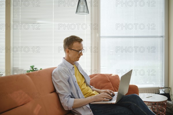 A retired man sits comfortably on a couch and writes his memoirs on laptop in a well-lit living room. Sunlight filters through the blinds, creating a warm and creative atmosphere