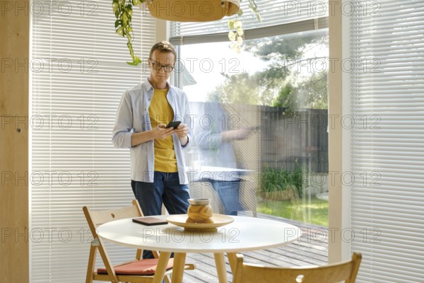 A middle-aged man stands in a bright, modern kitchen checking weather forecast at his smartphone. Sunlight filters through the blinds, illuminating the space