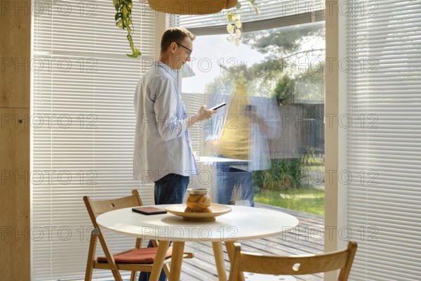 A man stands inside a stylish room, using his smartphone while looking out through a large glass window. Sunlight illuminates the space, and green plants are visible outside