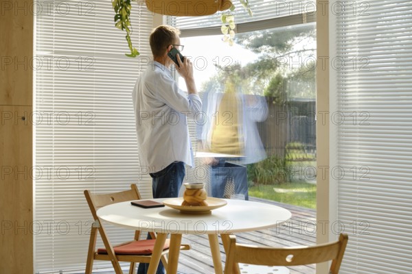 A middle-aged man stands near a large window, engaged in a phone conversation. Sunlight filters through the blinds, illuminating a contemporary dining area with a circular table and simple decor
