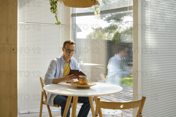 A man sits at a round wooden table in a modern kitchen, focused on his e-book. Sunlight filters through window blinds, creating a warm and inviting atmosphere as greenery decorates the space