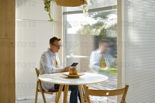 A middle-aged man sits at a small table, focused on reading an e-book in a well-lit room with large windows. Bright sunlight floods the space, illuminating the casual atmosphere and modern decor