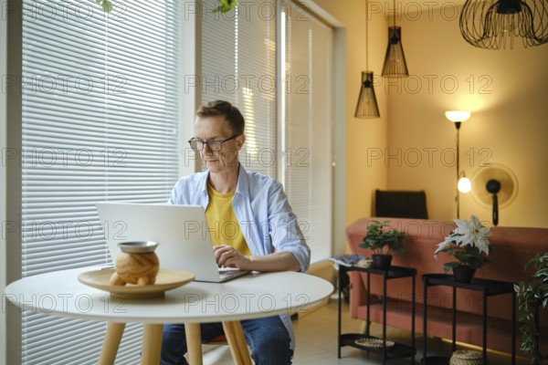 A freelancer is typing on a laptop at a round table in a modern home workspace. Natural light pours in through the blinds, illuminating the stylish decor and plants around