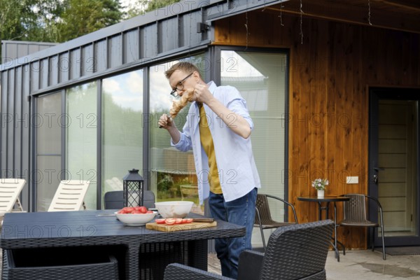 A man is enjoying a summer barbecue at a stylish wooden house. He is focused on grilling meat, surrounded by bowls of fresh ingredients and a pleasant outdoor setting