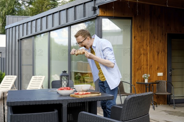 A middle-aged man stands at a patio table of tiny house and playfully bites marinated pork meat on skewer