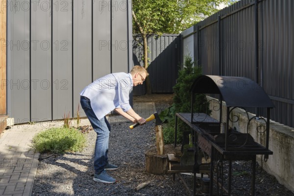 A middle-aged man is chopping a piece of wood in a well-maintained backyard garden. He is dressed casually in a light blue shirt and jeans while surrounded by greenery and a gray fence