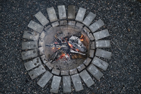 Bright flames dance among burning logs in a circular stone fire pit during early evening. The warm glow reflects off the dark gravel surrounding the fire, creating a cozy atmosphere under twilight