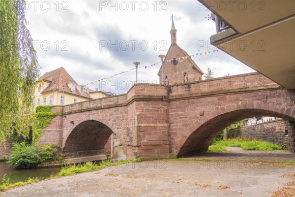 Large stone bridge with a church in the background under a cloudy sky, decorated with colourful flags, small town of Perle Calw, Black Forest, Germany