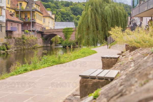Paved path along a river with stone wall and benches, a bridge in the background, small town of Perle Calw, Black Forest, Germany