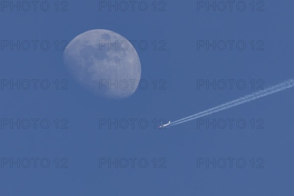 Boeing 737 jet passenger aircraft of Norweign Air with a vapour trial or contrail behind flying in a blue sky passing by the moon, England, United Kingdom