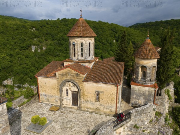 Close-up view of a church with tiled roofs and stone wall in front of wooded hills, aerial view, Motsameta Monastery, near Kutaisi, Imereti region, Georgia