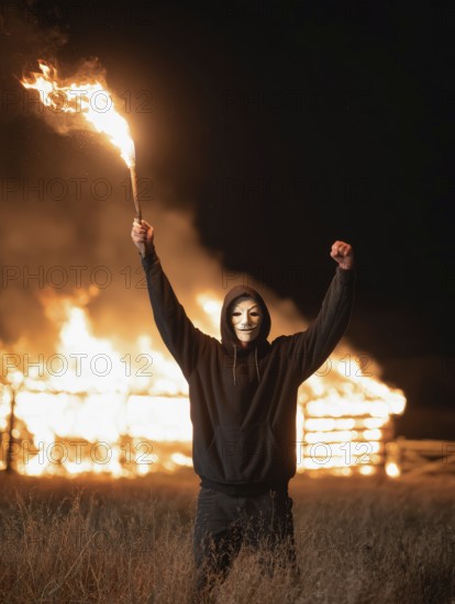 Maskierte Person hebt Fackel vor brennendem Strohlager in der Nacht, Brandstiftung, Brandleger, Feuerteufel, Pyromane, Pyromanie, Kriminalität, dunkle und bedrohliche Atmosphäre, AI generiert, AI generated