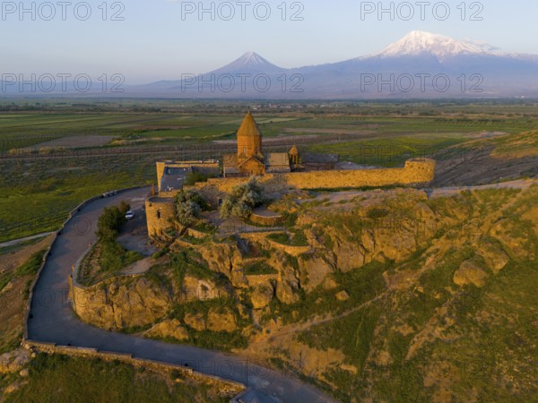 Historic monastery on a rocky hill with a wide view of mountains and plains in the warm light of sunrise, aerial view, Chor Wirap monastery, Chor Virap, deep dungeon, Ararat and Little Ararat in the background, located in Turkey, Ararat province, Armenia