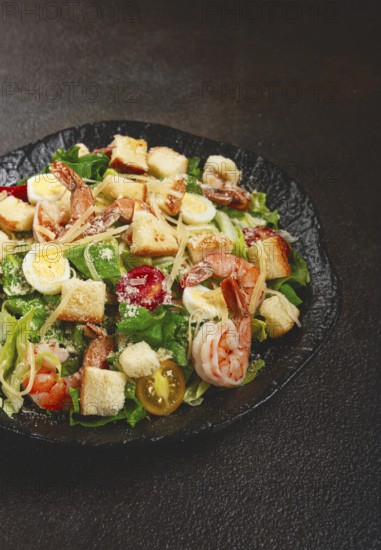Caesar salad with tiger prawns, shrimp salad, lettuce leaves, quail eggs, cherry tomatoes, parmesan cheese, on a black plate