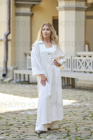 A middle-aged woman in a flowing white robe stands confidently outside a vintage architectural structure. The sun casts a warm glow, highlighting her relaxed yet poised demeanor as she showcases her outfit