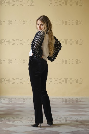 A middle-aged woman stands confidently with her back turned, showcasing a chic black and white outfit. The setting features a clean, light-colored wall and patterned tiles that enhance her fashionable presence