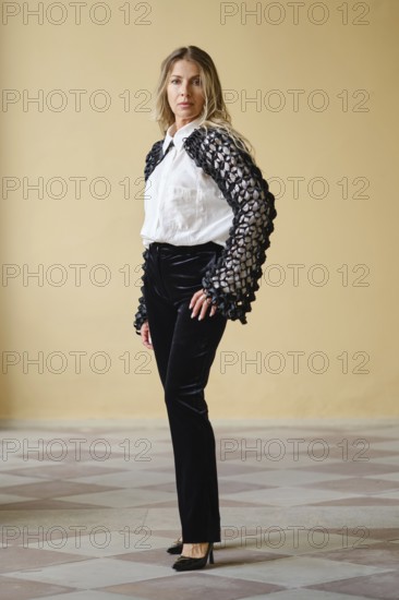 Brightly lit indoor location displays a middle-aged fashion model standing confidently. She wears a white shirt with distinctive, textured black sleeves paired with sleek black trousers