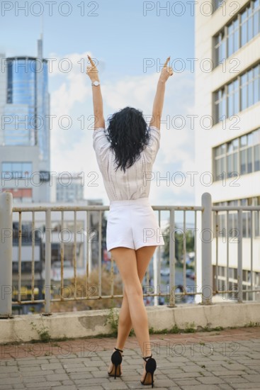 A woman stands outdoors in a vibrant urban setting with arms lifted to the sky. Dressed in a stylish outfit, she is enjoying a sunny afternoon surrounded by modern buildings