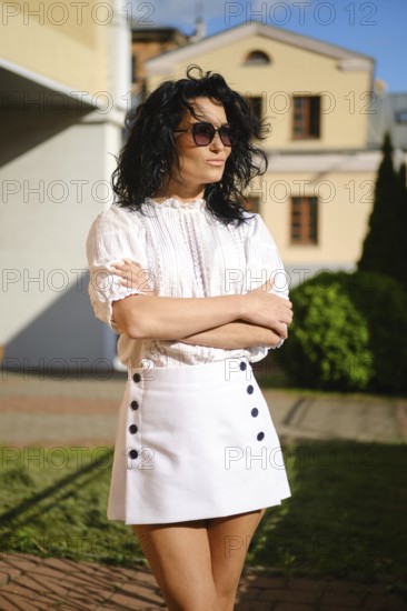 A woman with dark curly hair poses with crossed arms in a fashionable white outfit featuring a blouse and a high-waisted skirt with buttons