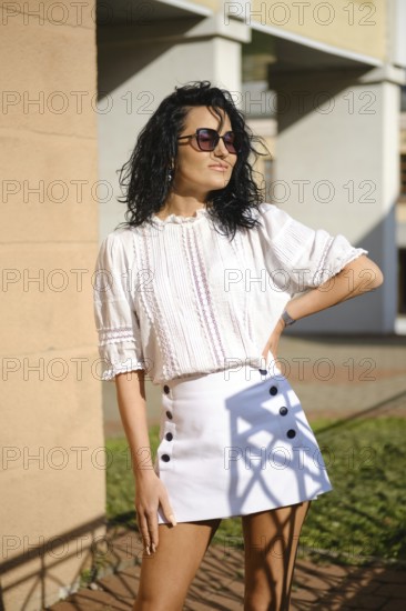 A woman with curly hair stands confidently in a fashionable outfit featuring a white blouse and a chic skirt with buttons. Sunlight casts playful shadows in the background, enhancing her look