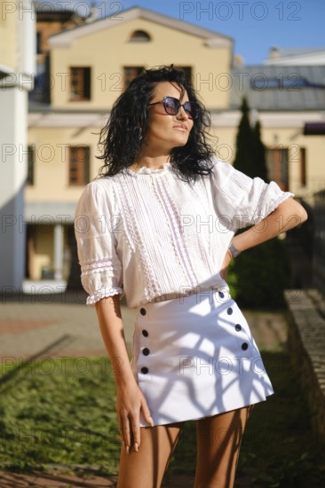 Bright sunlight highlights a woman posing confidently in a chic white blouse and skirt. She stands in a green area near a historic building, showcasing her sense of fashion and attitude
