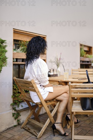 A woman with curly hair sits at a wooden table in a rustic outdoor cafe. She wears a white blouse and mini skirt pairing with high heels, relaxing while sipping a drink on a sunny day among greenery