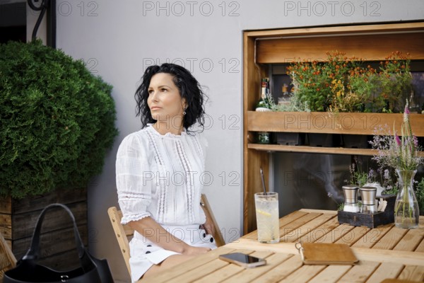 A woman with curly black hair sits at a wooden table in an outdoor cafe, gazing thoughtfully in a white outfit. She enjoys a cold drink while surrounded by greenery and a warm ambiance on a sunny day