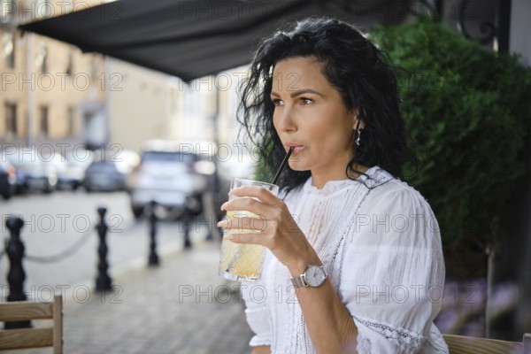 A woman sits at a sidewalk cafe, sipping a cold lemonade through a straw. She wears a white blouse and has long dark hair, while casually observing the city life around her