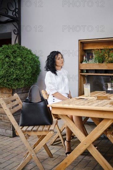 A woman with curly hair relaxes at a wooden table in an outdoor cafe, dressed in a fashionable white blouse. She enjoys a cold drink under the warm sun, surrounded by greenery and stylish decor