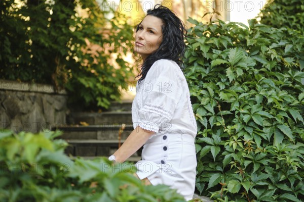 A woman with curly hair is seated gracefully on stone steps surrounded by vibrant green plants. She wears a stylish outfit and gazes thoughtfully into the distance on a sunny day