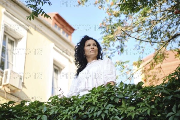 A woman with dark, wavy hair gazes thoughtfully while surrounded by lush green leaves in a charming courtyard. The sunlight highlights her features and the beautiful architecture behind her