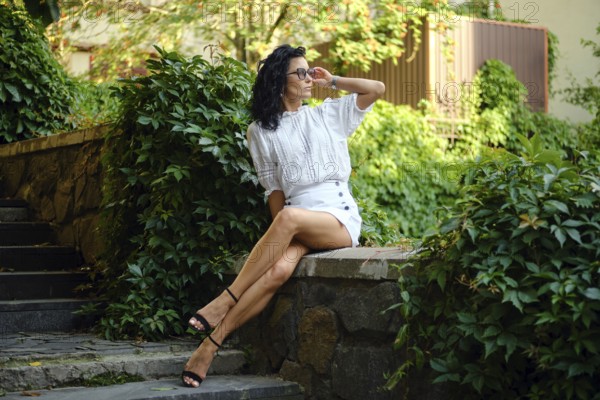A woman with curly hair is seated on a stone wall, wearing a stylish white outfit and sunglasses. Lush green foliage surrounds her, creating a vibrant atmosphere on a sunny day